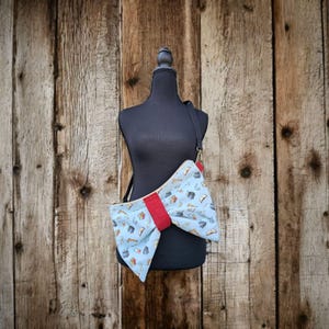 May include: A light blue crossbody bag with a bow design. The bag features a red center and a pattern of books and other objects. The bag is displayed on a black mannequin against a wooden background.