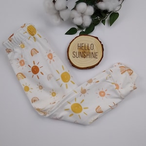 May include: White baby leggings with a sun and rainbow pattern. The leggings have a yellow, orange, and brown color scheme. The text "HELLO SUNSHINE" is on a wooden sign.