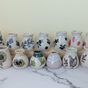 May include: A collection of thirteen small ceramic vases with a speckled cream finish. The vases feature various hand-painted designs including blueberries, sunflowers, strawberries, peaches, ginkgo leaves, and chickens in black and color.