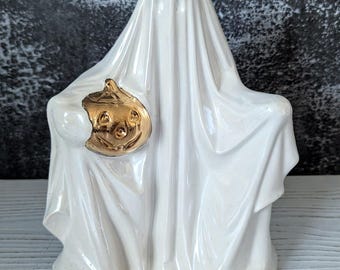 Puede incluir: Figurita de fantasma de cerámica blanca sosteniendo una calabaza dorada. El fantasma lleva una prenda similar a una sábana y tiene una expresión amigable.