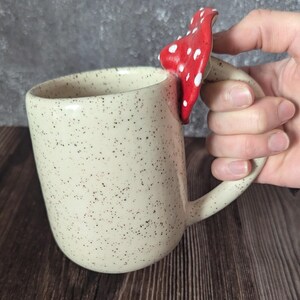 Peut inclure: Une tasse en c&eacute;ramique blanche avec un gla&ccedil;age brun mouchet&eacute; et une poign&eacute;e en forme de champignon rouge et blanc.