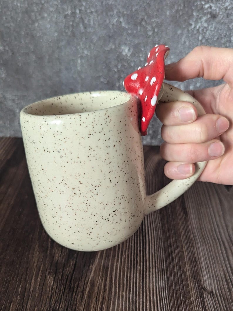 Peut inclure: Une tasse en c&eacute;ramique blanche avec un gla&ccedil;age brun mouchet&eacute; et une poign&eacute;e en forme de champignon rouge et blanc.