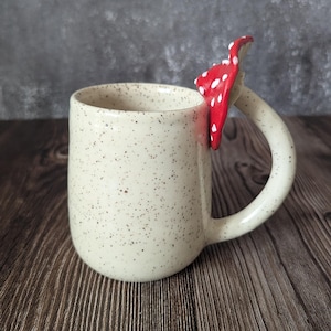 Peut inclure: Une tasse en c&eacute;ramique blanche avec une finition mouchet&eacute;e et une poign&eacute;e en forme de champignon rouge et blanc.
