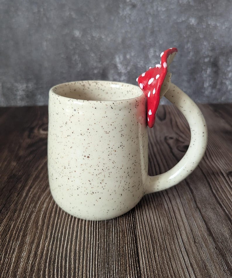 Peut inclure: Une tasse en c&eacute;ramique blanche avec une finition mouchet&eacute;e et une poign&eacute;e en forme de champignon rouge et blanc.