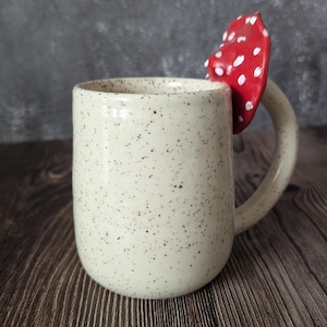 Peut inclure: Une tasse en c&eacute;ramique blanche avec des mouchetures brunes et une anse en forme de champignon rouge et blanc.