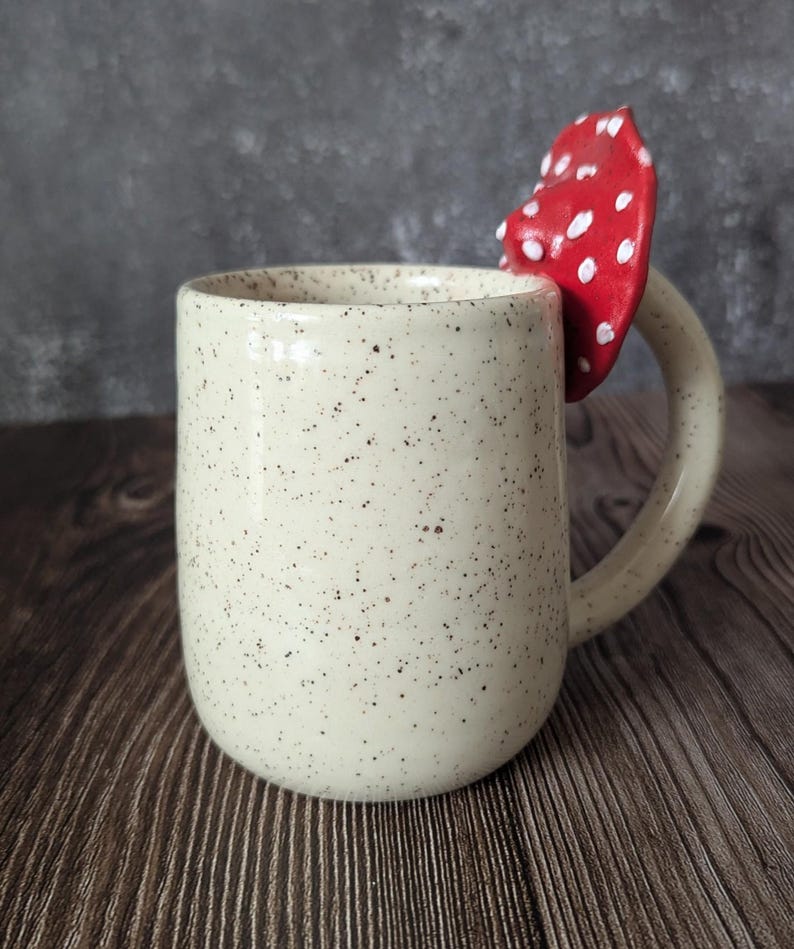 Peut inclure: Une tasse en c&eacute;ramique blanche avec des mouchetures brunes et une anse en forme de champignon rouge et blanc.