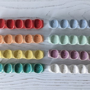 May include: Set of eight ceramic paint palettes in various colors, each with five round wells. The palettes are arranged in a row on a white wooden surface.