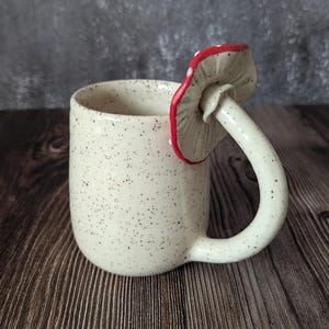 Peut inclure: Une tasse en c&eacute;ramique blanche avec une finition mouchet&eacute;e et une anse en forme de champignon. Le champignon est rouge avec des points blancs et a une tige blanche.