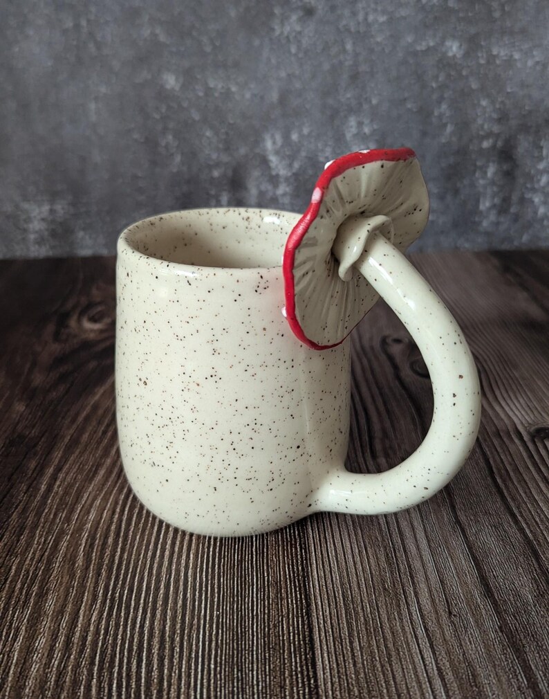 Peut inclure: Une tasse en c&eacute;ramique blanche avec une finition mouchet&eacute;e et une anse en forme de champignon. Le champignon est rouge avec des points blancs et a une tige blanche.