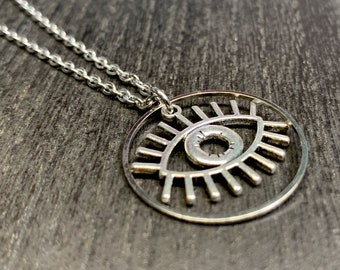 May include: A silver-colored necklace featuring a pendant with an eye design. The pendant is a circular frame with an eye shape in the center, and is suspended from a delicate chain. The necklace is displayed on a dark wooden surface.