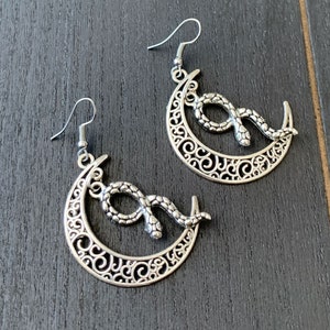May include: A pair of silver earrings featuring a crescent moon design with a snake wrapped around it. The earrings have a delicate, intricate design.