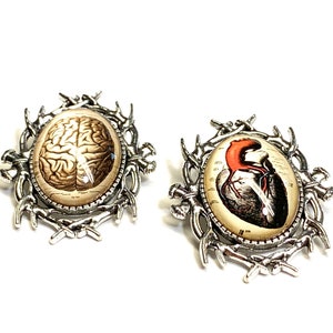 May include: Two silver-toned brooches with detailed, ornate frames. One features a vintage anatomical illustration of a human brain, the other a human heart.
