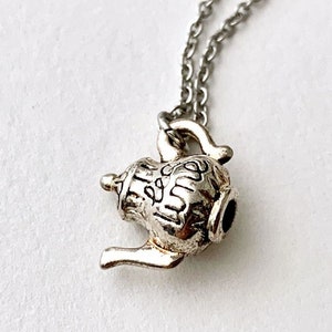 May include: A silver teapot charm necklace. The charm is detailed with a textured surface and the word "tea" inscribed on it.