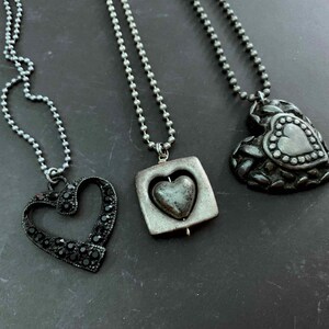 May include: Three heart-shaped necklaces on a dark surface. One is an open heart design with black stones, another has a heart within a square, and the third is a textured heart pendant. All have silver ball chains.