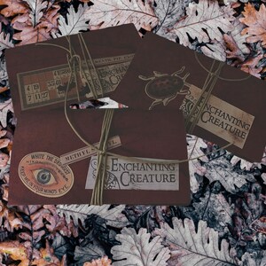 May include: Three vintage-style brown paper envelopes with white text and illustrations. The envelopes have the text "Enchanting Creature" and "White the Druggist" printed on them. One envelope has a drawing of a beetle, another has a drawing of an eye, and the third has a grid of numbers.