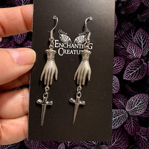 May include: Silver dangle earrings featuring a hand holding a dagger. The earrings are displayed on a black card with the text "Enchanting Creatu".