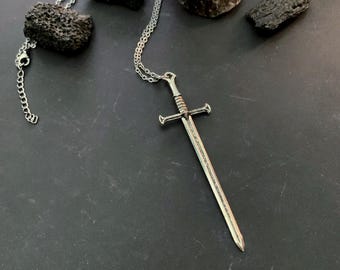 May include: A silver-toned sword pendant suspended from a delicate metal chain. The sword features a detailed hilt with a crossguard and a straight, pointed blade. The necklace is displayed alongside several dark, textured volcanic rocks.