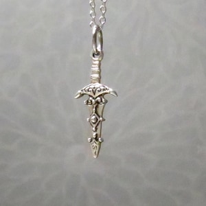 May include: A silver-colored pendant necklace featuring a detailed miniature sword. The sword pendant has an ornate hilt and blade, suspended from a delicate chain. The design is intricate, with decorative elements.