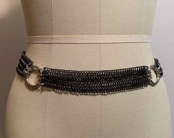 May include: A dark gray chainmail belt with two silver-colored circular clasps. The belt is composed of multiple rows of small, interlocking metal rings. The belt is displayed on a mannequin.