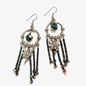 May include: Silver hoop earrings with iridescent crystal beads and a silver fairy charm hanging from each hoop. The earrings have a beaded fringe with iridescent crystal beads.