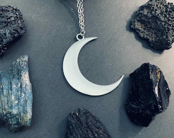 May include: A silver crescent moon pendant necklace. The pendant is a simple, polished crescent shape hanging from a delicate chain. The necklace is surrounded by dark, textured stones in shades of black and blue.