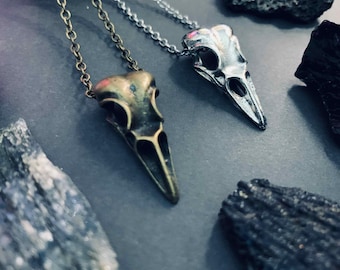 Op de afbeelding: Twee kettingen met raven schedel hangers. Eén is bronskleurig, de andere zilverkleurig, elk hangend aan een delicate ketting. De hangers zijn geplaatst tegen een donkergrijze achtergrond met ruwe, donkere stenen.