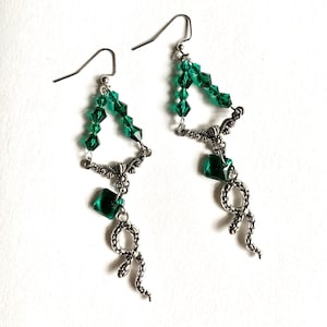 May include: A pair of dangle earrings with a green crystal triangle design and a snake charm. The earrings are made with silver-toned metal.