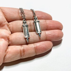 May include: Two silver syringe necklaces on a hand. The necklaces are silver and have a detailed design.