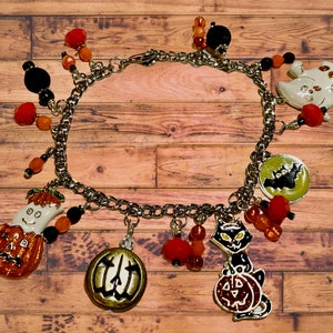 May include: A silver charm bracelet with Halloween-themed charms, including pumpkins, ghosts, bats, and a black cat. The charms are made of metal and have a shiny finish. The bracelet is adorned with orange and black beads.