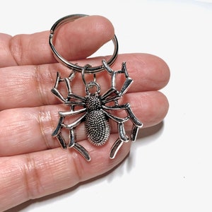 May include: Silver spider keychain with a detailed web design. The spider has a textured body and eight legs.
