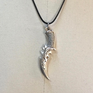 May include: Silver-colored dagger pendant on a black cord necklace. The dagger features a textured handle and ornate, curved blade. The pendant is suspended from a simple loop and hangs against a neutral background.