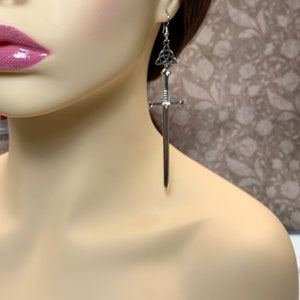 May include: Silver sword earrings with a Celtic knot charm. The sword is long and slender with a pointed tip. The earrings are dangling and have a silver hook.