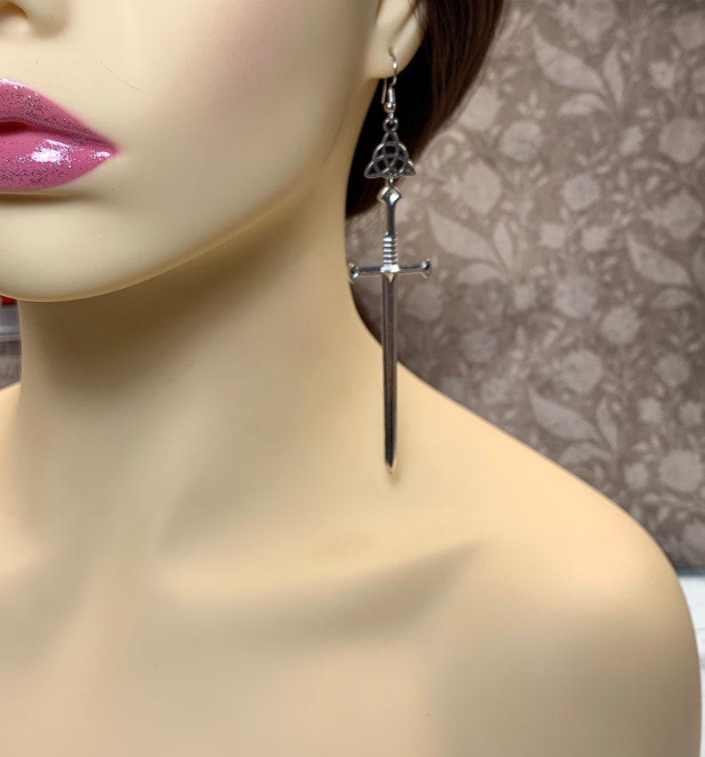 May include: Silver sword earrings with a Celtic knot charm. The sword is long and slender with a pointed tip. The earrings are dangling and have a silver hook.