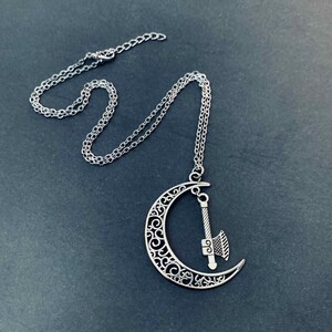 May include: A silver-toned necklace featuring a crescent moon pendant with intricate filigree cutouts. A small, detailed axe charm hangs from the center of the moon. The pendant is attached to a delicate silver-toned chain with a lobster claw clasp.