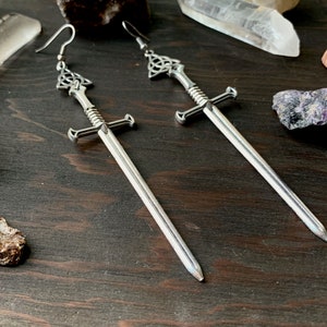May include: A pair of silver sword earrings with a Celtic knot design at the top of each sword. The earrings are hanging from a dark wooden surface.
