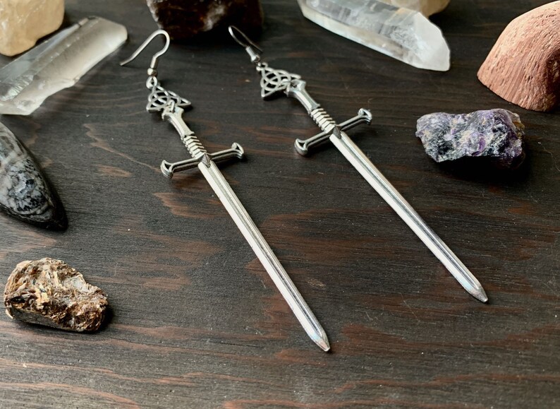 May include: A pair of silver sword earrings with a Celtic knot design at the top of each sword. The earrings are hanging from a dark wooden surface.