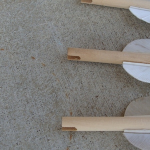 May include: Three wooden arrows with white fletching. The arrows are lying on a concrete surface.