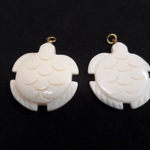 May include: Two white carved turtle pendants with gold accents. The turtles have detailed shell carvings and are ready to be strung on a necklace or bracelet.