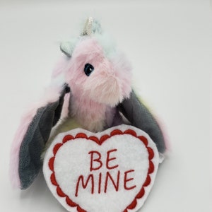 Puede incluir: Un unicornio de peluche rosa y azul sosteniendo una almohada blanca en forma de corazón con las palabras "BE MINE" bordadas en rojo.
