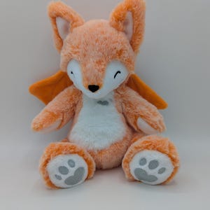 May include: A plush orange fox toy with white accents on the chest and paws. The fox has large ears, a black nose, and gray paw prints on its feet. It also has small orange wings.