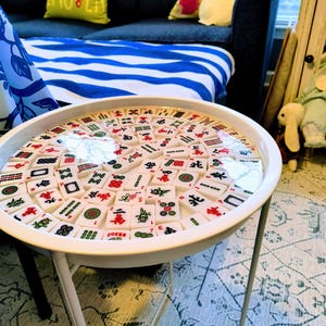 May include: A white, round side table with a tabletop inlaid with numerous Mahjong tiles. The tiles are white with red, green, and black symbols. The table has a simple, three-legged design and is set against a patterned rug.