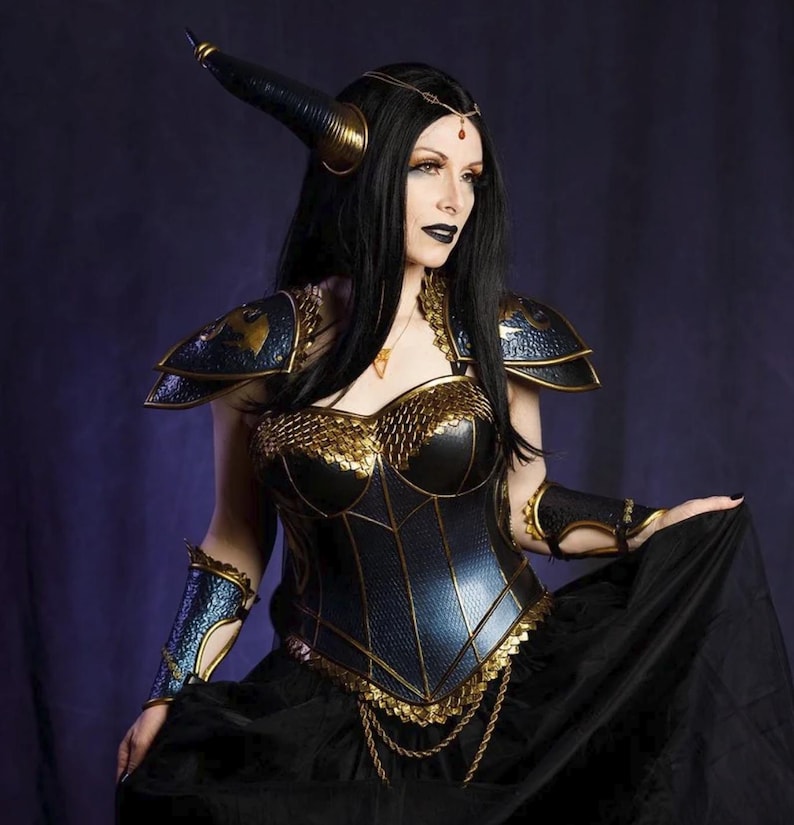 May include: A fantasy costume featuring a black and gold corset, shoulder armor, and arm cuffs. The costume includes a black skirt and a horned headpiece. The overall design has a medieval or gothic aesthetic.