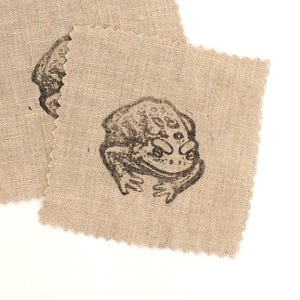 May include: Four beige fabric squares with a black frog illustration. Each square has a jagged edge. The frog design is centered on each square. The squares are arranged on a white background.