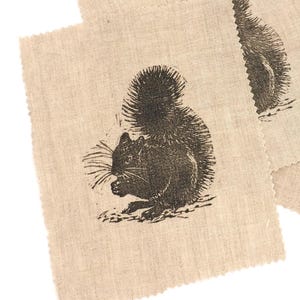 May include: Three beige fabric squares with a black squirrel illustration. The squirrel is depicted in a detailed, vintage style, holding a nut. The fabric has a natural, textured appearance with a raw, unfinished edge.