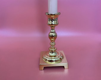 May include: A gold metal candle holder with a square base and a round top. The candle holder is holding a white candle.