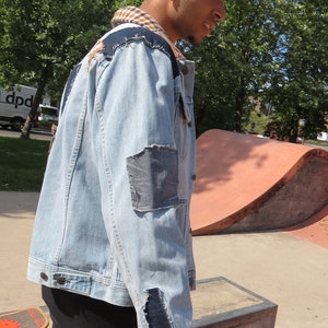 May include: A light blue denim jacket with several patches sewn on. The jacket has a frayed collar and a few rips in the fabric. The person wearing the jacket is standing in front of a skate ramp.