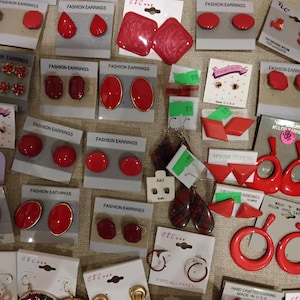 May include: A collection of various red fashion earrings displayed on individual white cardboard backing cards. The assortment includes circular, oval, square, diamond, and triangular shapes, some with gold-tone accents or plaid patterns.