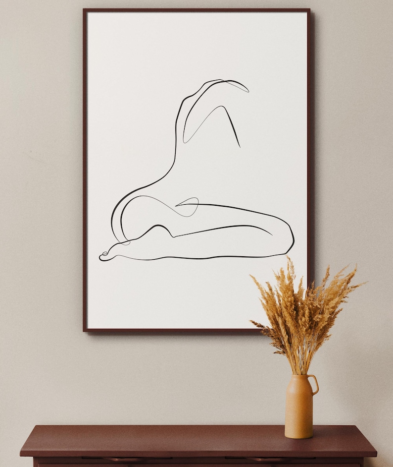 May include: A black and white line drawing of a person in a seated pose, framed in a brown frame and hanging on a light beige wall. A vase with dried wheat stalks is on a dark brown table in front of the artwork.