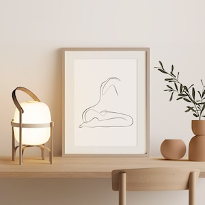 May include: A white table lamp with a wooden base and a white shade. The lamp is on a wooden table with a framed print of a line drawing of a person sitting. A potted plant is on the table to the right of the lamp.