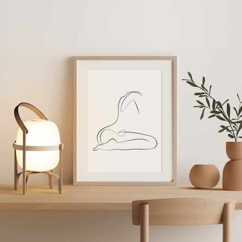 May include: A white table lamp with a wooden base and a white shade. The lamp is on a wooden table with a framed print of a line drawing of a person sitting. A potted plant is on the table to the right of the lamp.